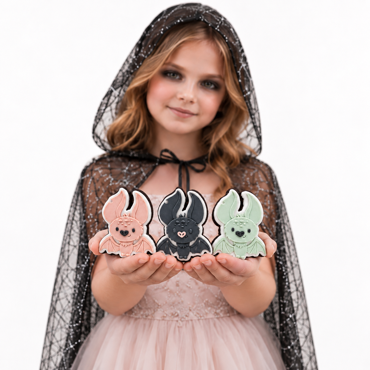 Girl in a black hooded cape presenting three Halloween bat sugar cookies.