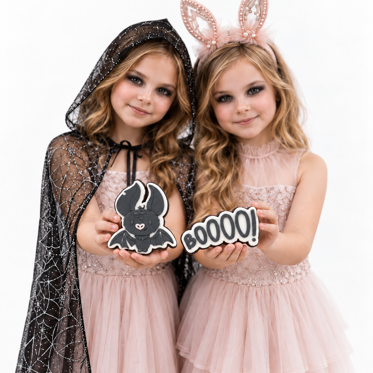 Two young girls in pink and black Halloween costumes holding 'BOOOOOO!' and a bat-shaped cookie, posing against a white background.