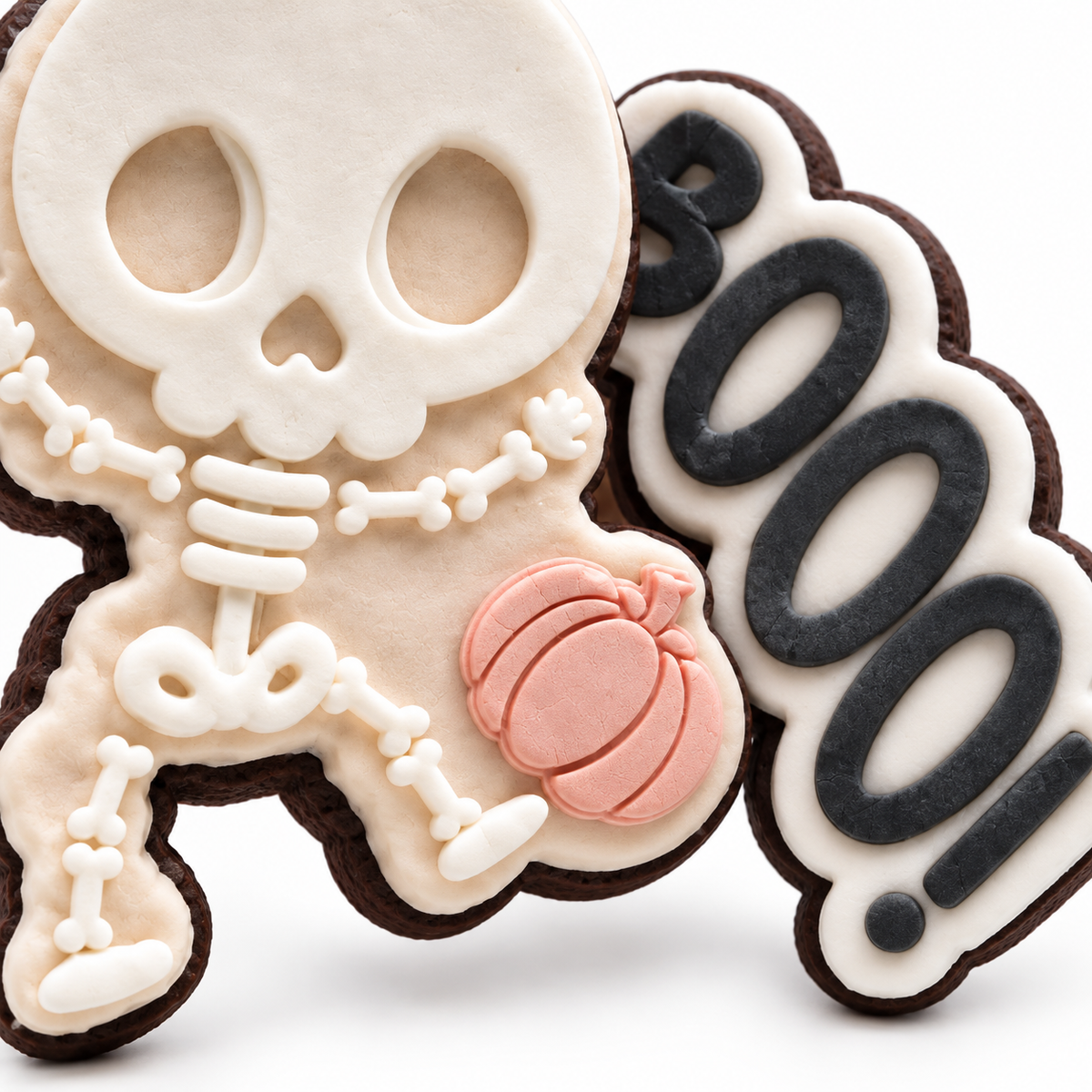 Close-up of a fondant skeleton Halloween cookie with a blush pumpkin beside a charcoal BOOOOO cookie.