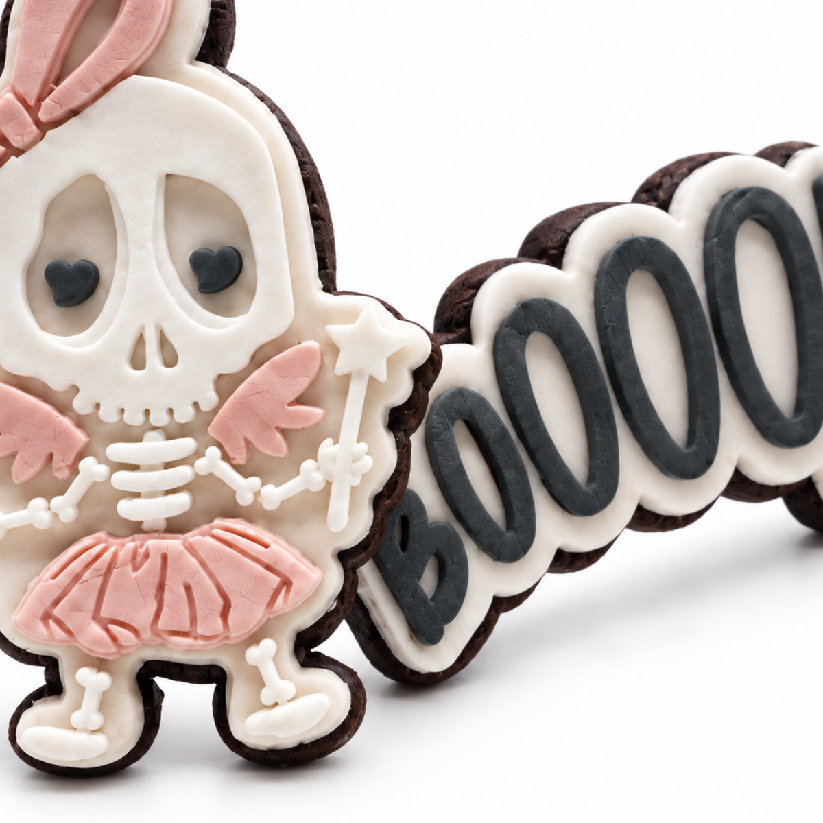 Close-up of a fondant skeleton girl Halloween cookie with a blush bow and tutu beside a charcoal BOOOOO cookie.