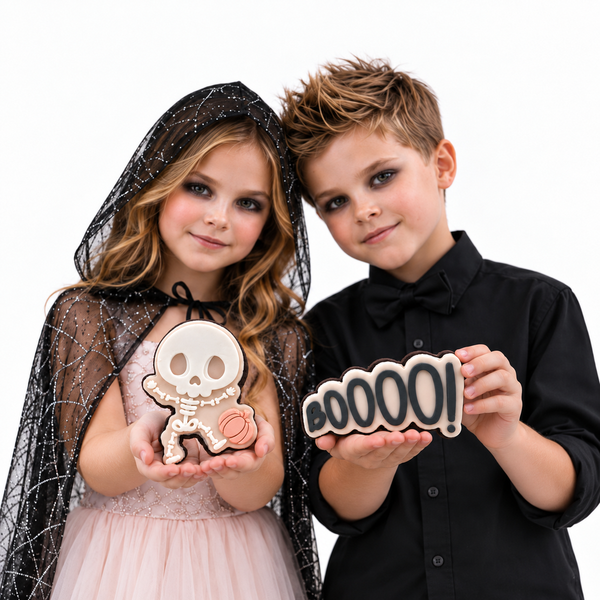 Girl in a spiderweb cape and boy in black presenting Little Bones and BOOOO Halloween cookies.