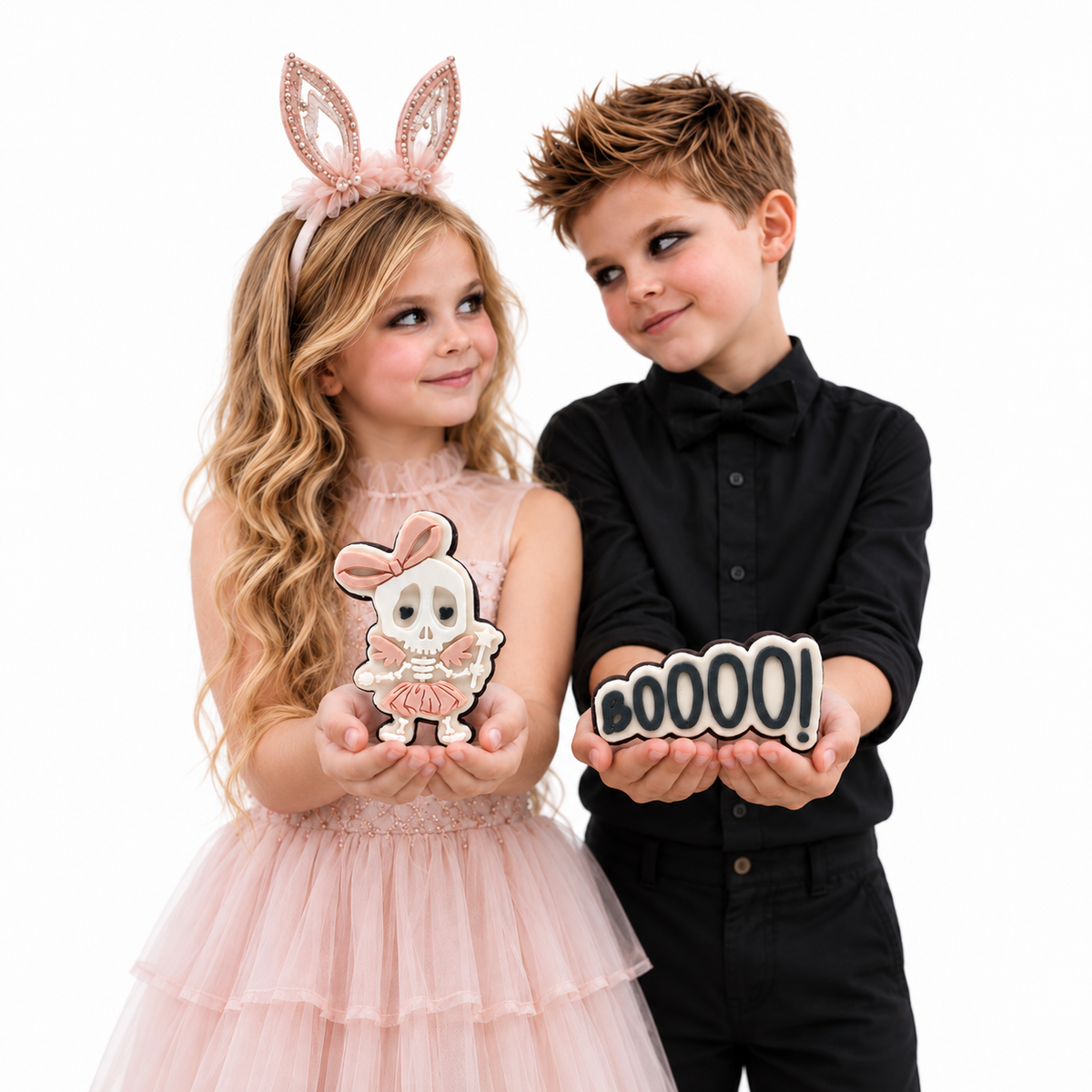 Girl in blush bunny ears and boy in black presenting skeleton ballerina and BOOOO Halloween cookies.
